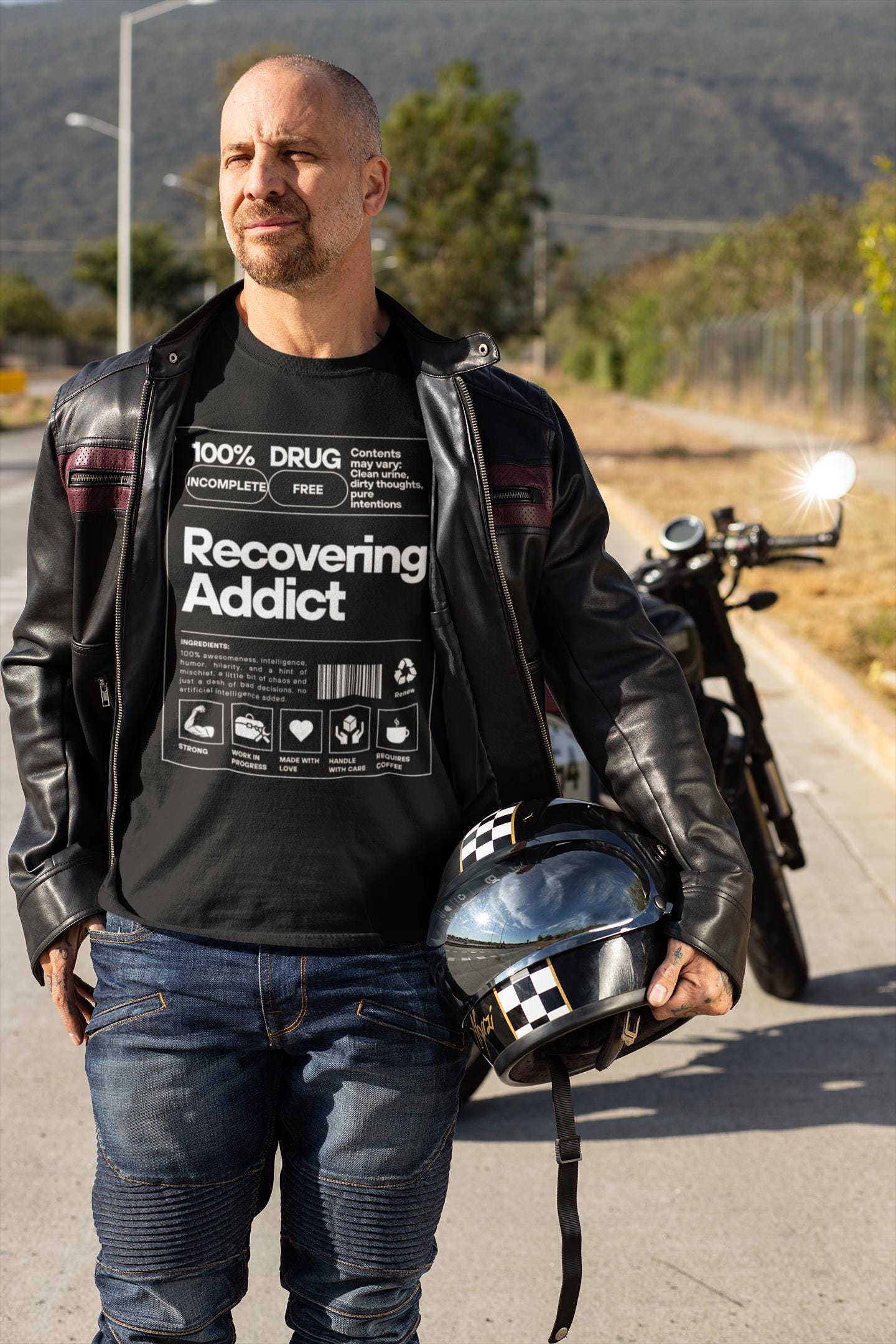 Recovering addict T-shirt worn by a man holding a helmet in the foreground and a motorcycle in the background.