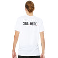 Person wearing a white t-shirt with 'STILL HERE' text on the back, standing against a white background